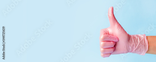 Closeup of female hand in pink cosmetic medical hygiene pharmacy gloves showing thumbs up sign against pastel blue background, copy space, minimal concept