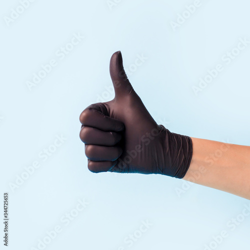 Closeup of female hand in black cosmetic medical hygiene pharmacy gloves showing thumbs up sign against pastel blue background, copy space, minimal concept