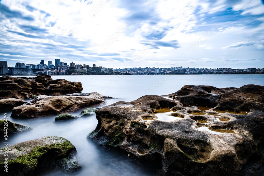 Long exposure on a partly cloudy afternoon on Sydney Harbour with nice rocks in the foreground the soft waves crashing on the shore and the beautiful harbour foreshore as a backdrop