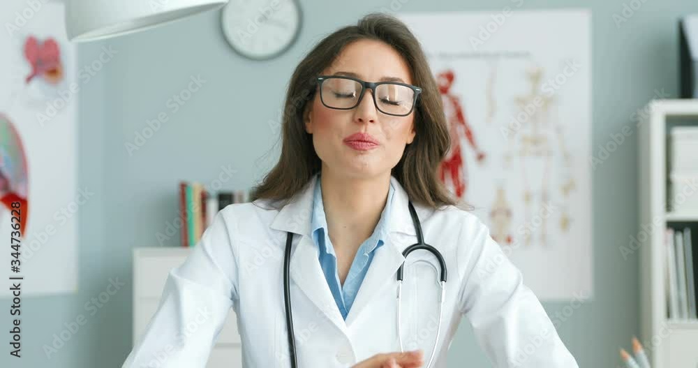 Portrait of Caucasian young female doctor in glasses and white gown sitting at table and talking to camera as videoblogging about health. Woman medic bloger speaking on healthcare Medical work concept Portrait of Caucasian young female doctor in glasses and white gown sitting at table and talking to camera as videoblogging about health. Woman medic bloger speaking on healthcare Medical work concept