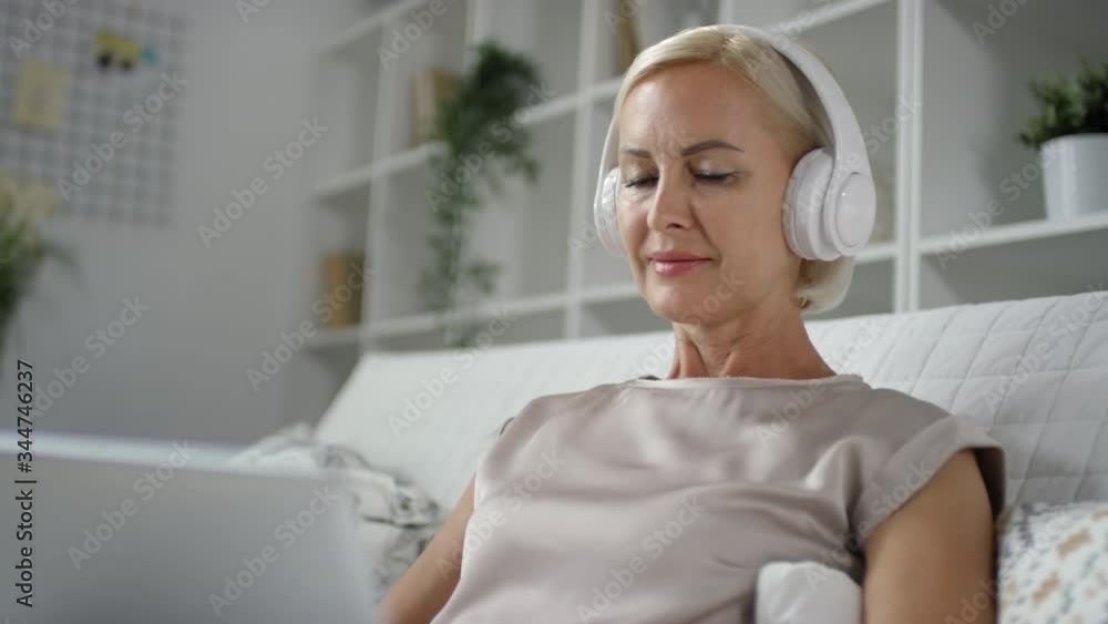 Smiling mature woman sitting on sofa at home, listening to music with headphones and surfing the Internet on laptop Smiling mature woman sitting on sofa at home, listening to music with headphones and surfing the Internet on laptop