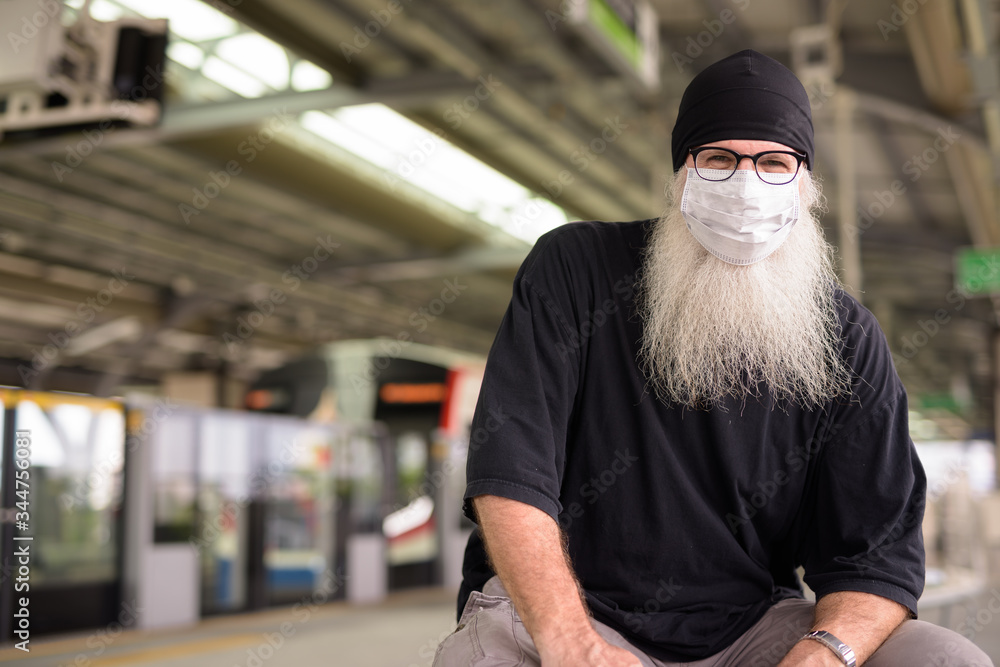 Mature bearded hipster man with mask for protection from corona virus outbreak sitting at train station