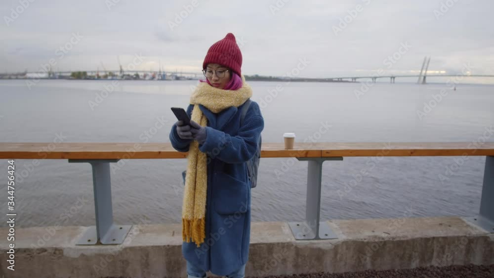 Zoom out of young Asian female tourist with backpack standing on city riverside with cup of coffee, looking at bridge view and taking selfie with smartphone while having walk on autumn day Zoom out of young Asian female tourist with backpack standing on city riverside with cup of coffee, looking at bridge view and taking selfie with smartphone while having walk on autumn day