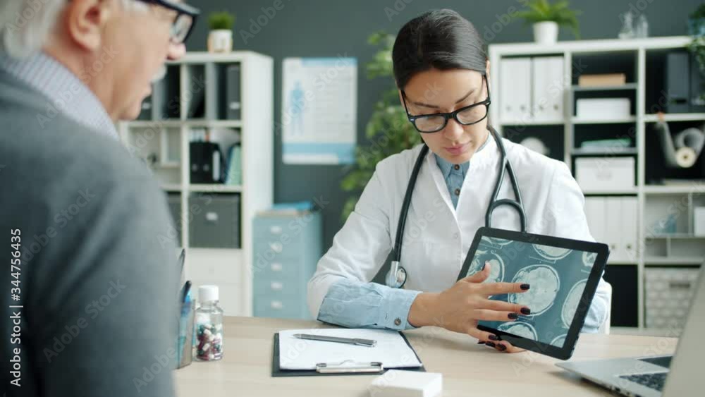 Young lady doctor is showing tablet screen with MRI results to senior patient in hospital office touching screen and talking discussing disease and treatment. Young lady doctor is showing tablet screen with MRI results to senior patient in hospital office touching screen and talking discussing disease and treatment.