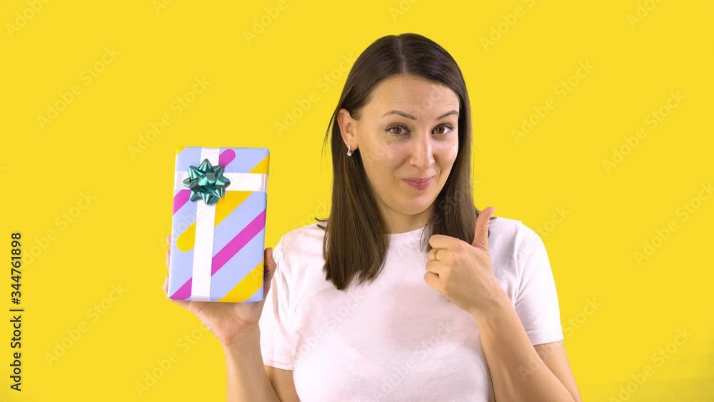 Young Woman in White T-shirt Holding Beautiful Box with a Gift and Showing Thumb Up. Back Yellow Background. Holiday Mood. Young Woman in White T-shirt Holding Beautiful Box with a Gift and Showing Thumb Up. Back Yellow Background. Holiday Mood.