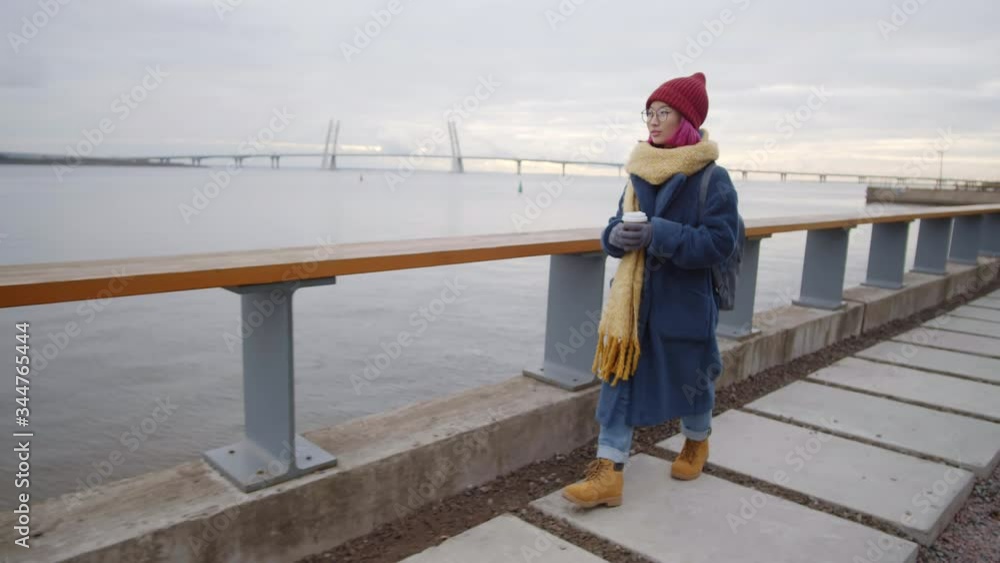 Young Asian female tourist with backpack holding coffee cup and looking at beautiful city view while walking along urban riverside on autumn day Young Asian female tourist with backpack holding coffee cup and looking at beautiful city view while walking along urban riverside on autumn day