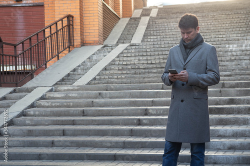 elegant man in long overcoat reads information using digital technology and standing on high steps. Concept fashion city technology