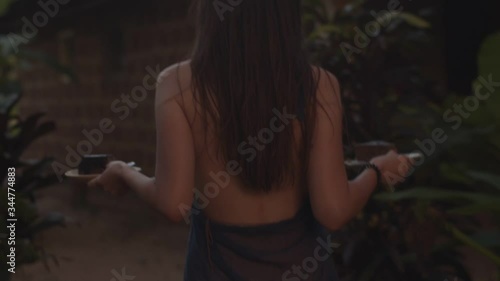 Back view of unrecognizable woman carrying plates with food outdoors natural twilight light. Faceless elegant girl in dress with no back going outside slow motion. Female beauty gracefulness concept