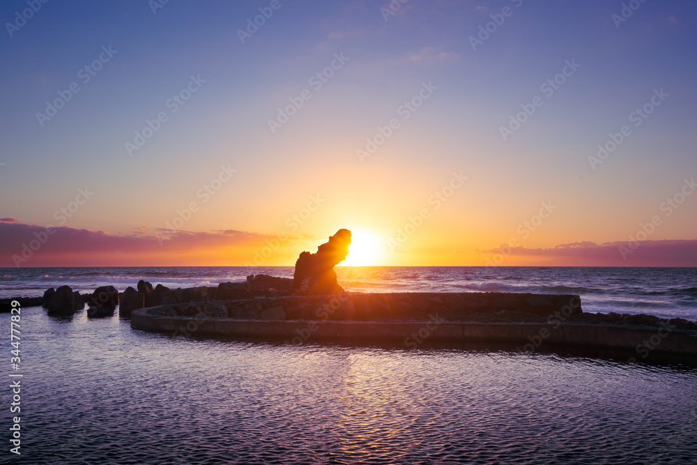 Sunset view on rock with reflecting sun rays against beautiful sea and gentle violet sky. Romantic sunset sea background.