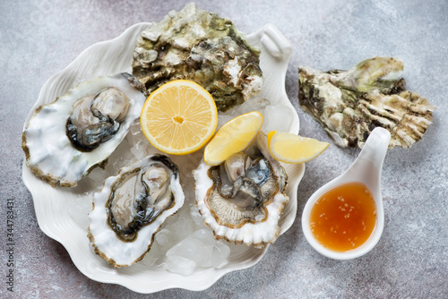 White shell-shaped plate with opened oysters on ice and lemon, horizontal shot on a beige stone background, selective focus
