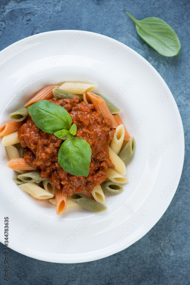 White plate with three-colored penne and bolognese sauce, vertical shot on a blue stone background, above view