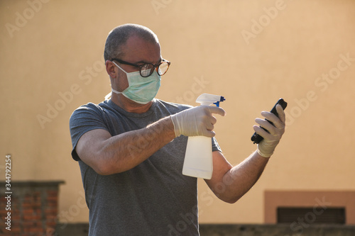 Man in the protective mask and gloves cleans a mobile phone using bottle with desinfectant in spray photo