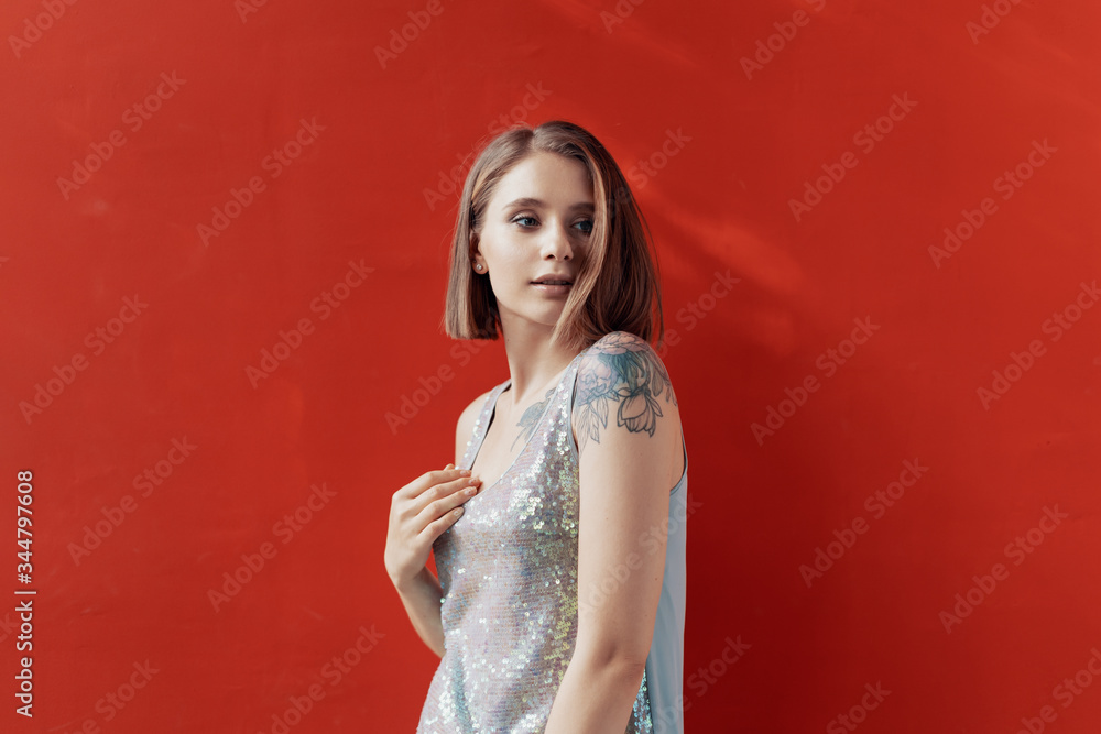 Cheerful young girl posing in front of a red wall. Natural light. Tattoo on left shoulder. Dressed in a shiny dress Cheerful young girl posing in front of a red wall. Natural light. Tattoo on left shoulder. Dressed in a shiny dress