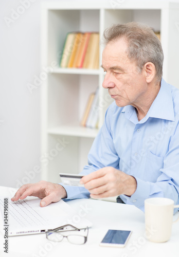 Male pensioner working makes purchases at home using laptop and credit card.