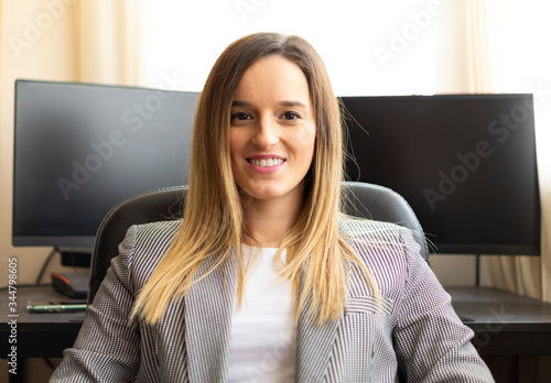 Young attractive businesswoman smiling at the camera while sitting in a executive chair with computers on the background.Woman telecommuting from home. Entrepreneur woman. Successful woman.