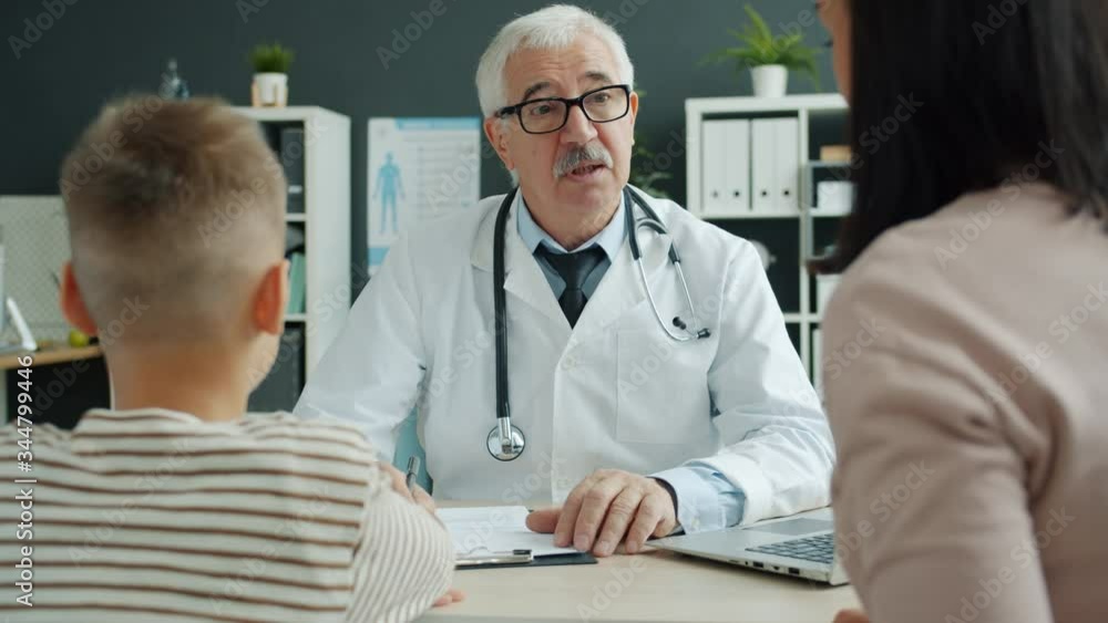 Experienced paediatrist talking to mother and child writing in medical record in clinic office sitting at desk in modern office. People, medicine and healthcare concept. Experienced paediatrist talking to mother and child writing in medical record in clinic office sitting at desk in modern office. People, medicine and healthcare concept.
