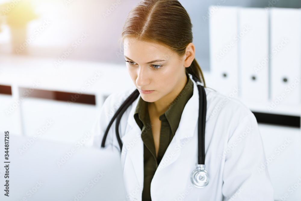 Woman-doctor typing on pc computer while sitting at the desk in sunny hospital office. Data in medicine and healthcare Woman-doctor typing on pc computer while sitting at the desk in sunny hospital office. Data in medicine and healthcare