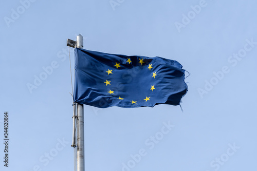 Deteriorated European Union flag blowing in the wind in direct sunlight towards clear blue sky, torn textile material 