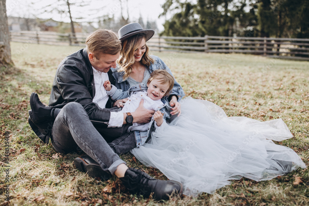 Fototapeta premium Stylish family in the autumn forest. A guy in a leather jacket and a young girl in a gray-blue dress play with his son in a meadow of a pine forest in the mountains against the background of a fence 