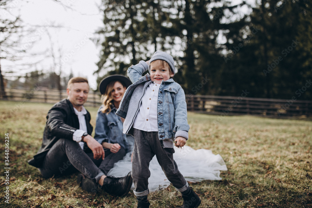 Stylish family in the autumn forest. A guy in a leather jacket and a young girl in a gray-blue dress play with his son in a meadow of a pine forest in the mountains against the background of a fence Stylish family in the autumn forest. A guy in a leather jacket and a young girl in a gray-blue dress play with his son in a meadow of a pine forest in the mountains against the background of a fence