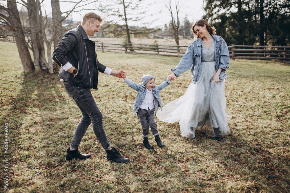 Fototapeta premium Stylish family in the autumn forest. A guy in a leather jacket and a young girl in a gray-blue dress play with his son in a meadow of a pine forest in the mountains against the background of a fence