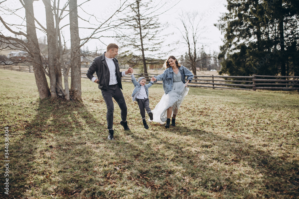 Fototapeta premium Stylish family in the autumn forest. A guy in a leather jacket and a young girl in a gray-blue dress play with his son in a meadow of a pine forest in the mountains against the background of a fence 