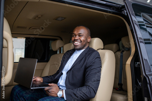 African successful businessman working at a laptop with a grin sitting in the beige interior of a large car