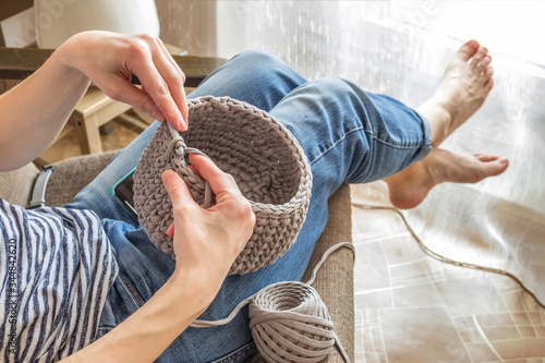 a young girl is sitting at home in quarantine in a chair and crocheting a basket made of gray knitted cotton yarn close up in blue jeans and a striped t shirt, faceless, anonymity