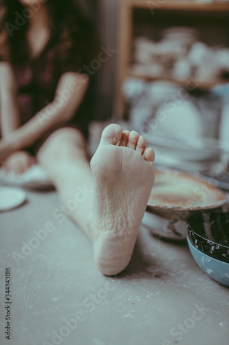 A close-up of barefoot girl sitting on the concrete floor. A bare hill stained with white plaster. A potterry girl making ceramics tableware. A ceramist in her workshop. A close view of hee and toes. photo