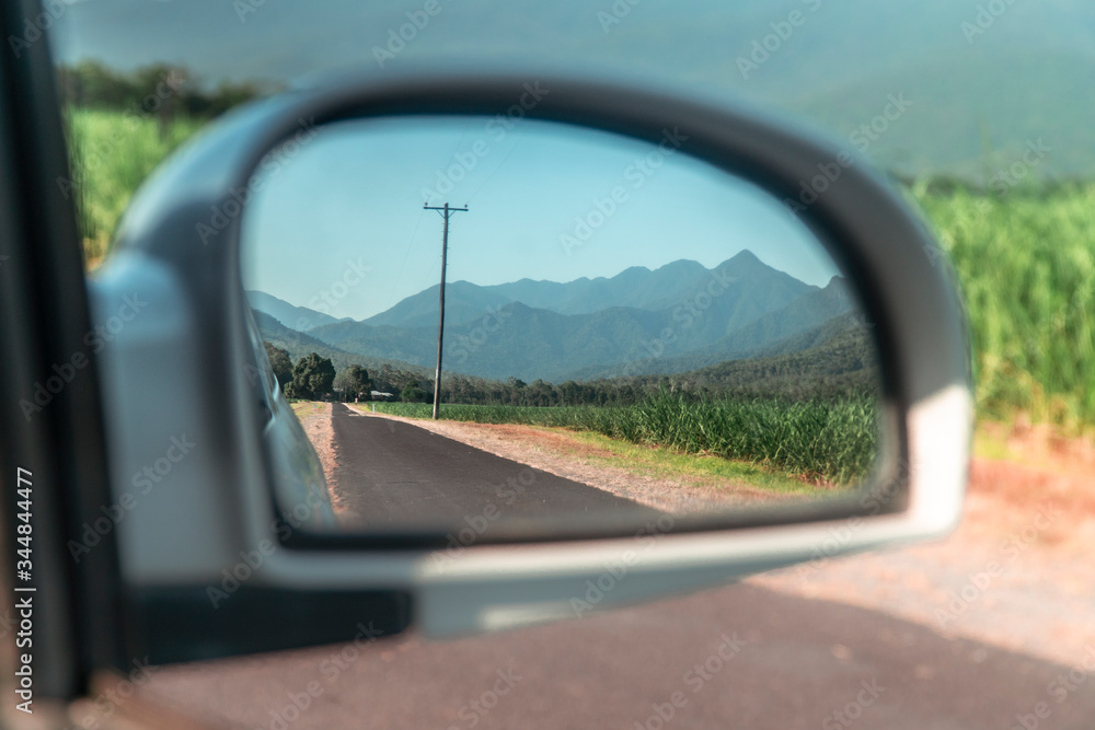 Rearview car mirror reflection of Beautiful mountain landscape with sugar cane fields foreground. Road, fields, trees, green forest, farm, mountains, blue sky & road. Walsh's Pyramid, Cairns Australia Rearview car mirror reflection of Beautiful mountain landscape with sugar cane fields foreground. Road, fields, trees, green forest, farm, mountains, blue sky & road. Walsh's Pyramid, Cairns Australia
