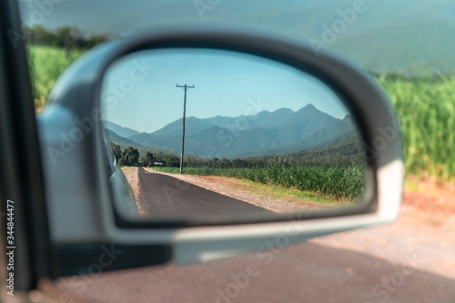 Rearview car mirror reflection of Beautiful mountain landscape with sugar cane fields foreground. Road, fields, trees, green forest, farm, mountains, blue sky & road. Walsh's Pyramid, Cairns Australia © Jam Travels