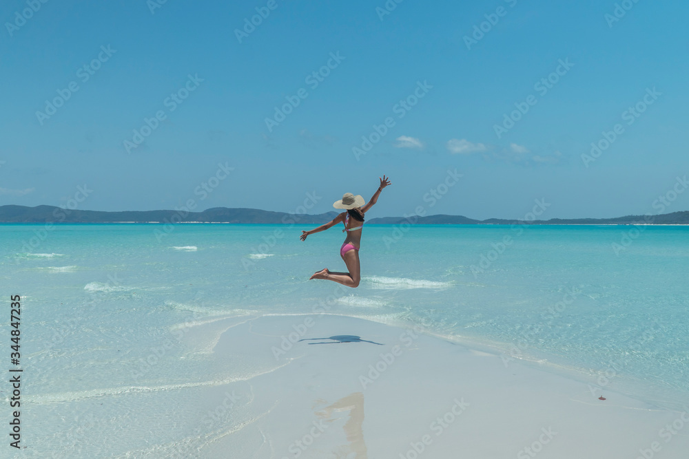 Woman jumping on blue beach. Tourist on Whitsundays beach, white sand, in pink bikini & hat, with aqua turquoise ocean. Travel, holiday, vacation, paradise, exotic. Whitsundays Islands, Australia.