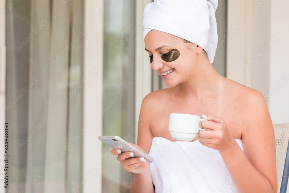 Wrapped in towel after shower woman with eye patches is drinking tea or coffe at the table and reading news or messages at the hotel terrace. Girl is enjoying her vacation Wrapped in towel after shower woman with eye patches is drinking tea or coffe at the table and reading news or messages at the hotel terrace. Girl is enjoying her vacation