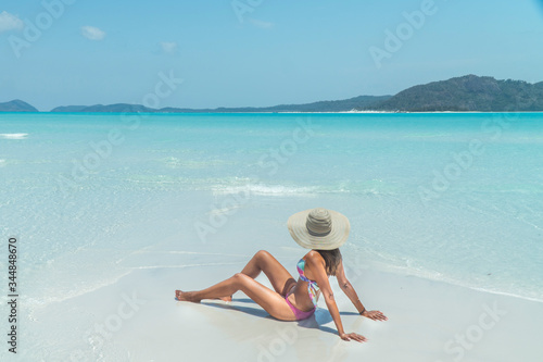 Woman sitting on paradise beach. Tourist on Whitsundays beach, white sand, in pink bikini & hat, with aqua blue turquoise ocean. Travel, holiday, vacation, exotic. Whitsundays Islands, Australia.