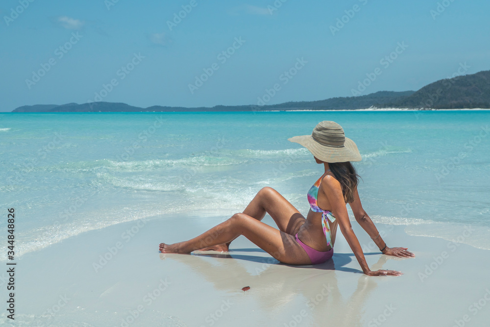 Woman sitting on paradise beach. Tourist on Whitsundays beach, white sand, in pink bikini & hat, with aqua blue turquoise ocean. Travel, holiday, vacation, exotic. Whitsundays Islands, Australia.