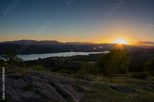 The setting sun creates a starburst effect as it sets below a mountain peak at the end of a large lake. Rocky and green terrain lead the viewer towards the lake and the setting sun. 