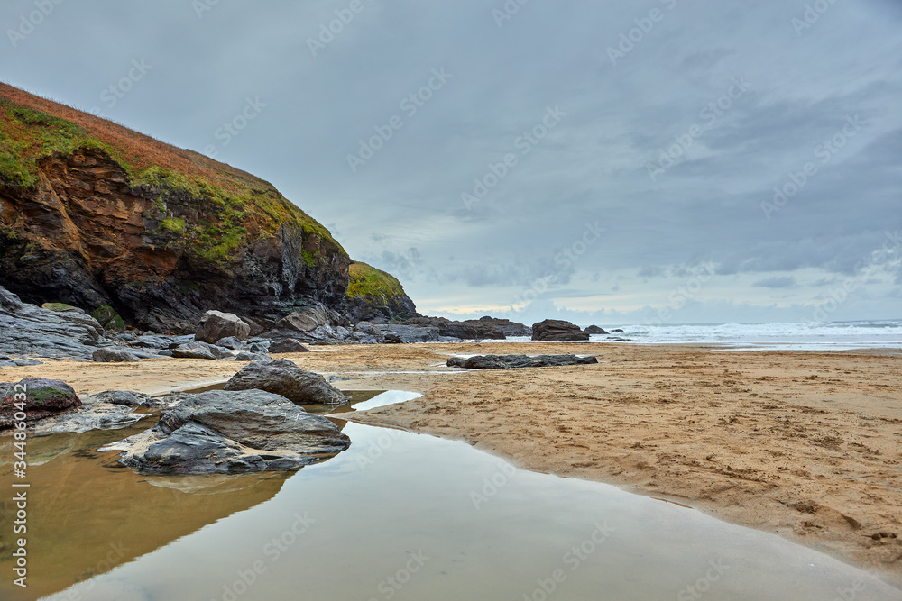 Image of Poldhu Beach in Cornwall with rocks, waves, sand, water in the foreground and cloudy sky. Image of Poldhu Beach in Cornwall with rocks, waves, sand, water in the foreground and cloudy sky.