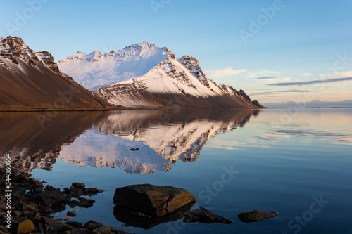 A snow capped mountain perfectly reflected in still blue water with rocks and grass in the foreground.