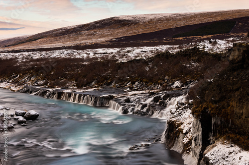 A turquoise blue river flows through a snowy autumnal landscape with water cascading over lava rock formations.