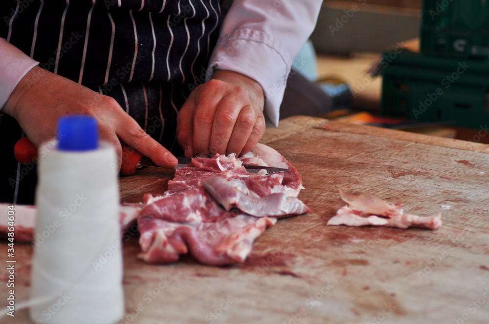 A man wearing an apron fillets a piece of meat using a knife on a chopping board. A spindle of string is out of focus in the foreground.
