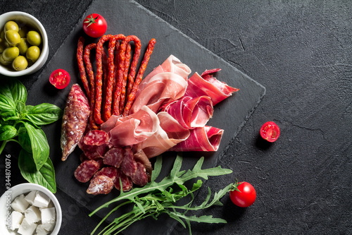 Tapas concept. Prosciutto, sausage chorizo, olives, cherry tomato, arugula, basil and cheese on a black slate board. Antipasti concept.