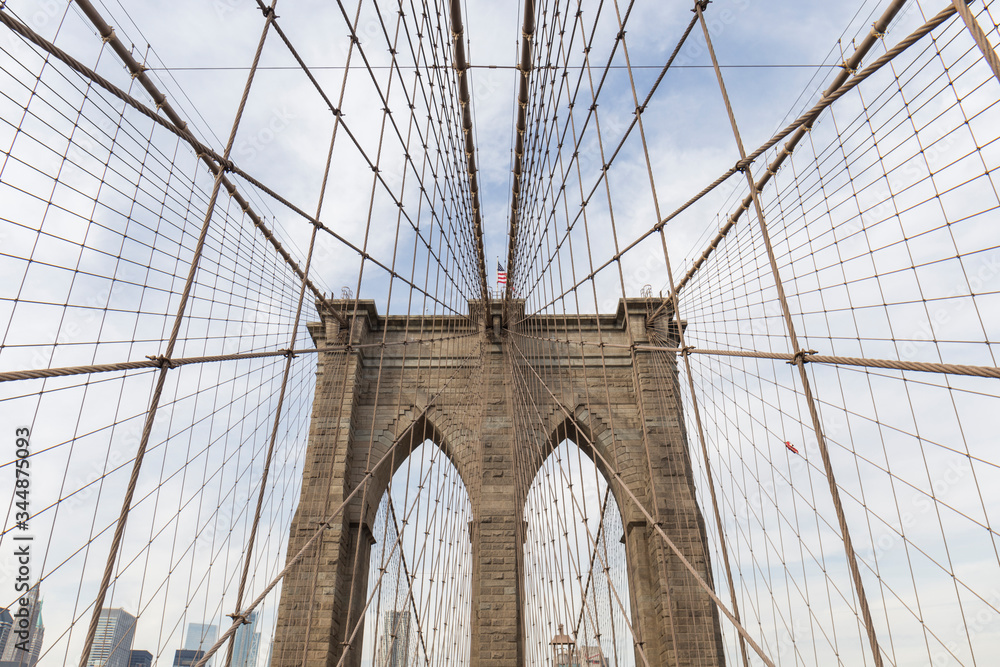 Fototapeta premium Brooklyn Bridge, zdjęcie z wnętrza mostu, perspektywa jednego z łuków z kablami tworzącymi geometryczne kształty na niebie, pośrodku flaga USA, zdjęcia Nowego Jorku