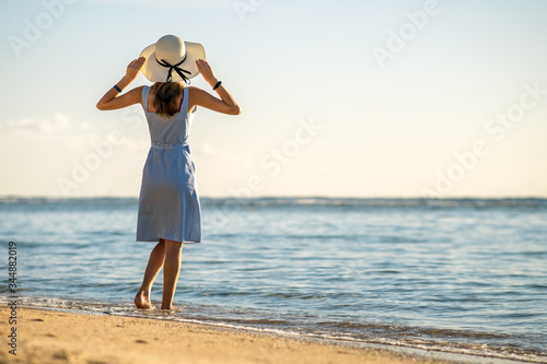 Young woman in straw hat and a dress standing alone on empty sand beach at sea shore. Lonely tourist girl looking at horizon over calm ocean surface on vacation trip.