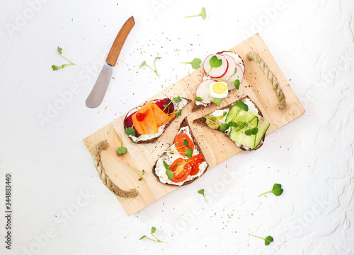 Rye bread sandwiches with cream cheese, vegetables and microgreen on wooden tray with butter spreader. Vegetarian tapas with cucumber, tomatoes, quail eggs, radish, carrot, beet, olives 