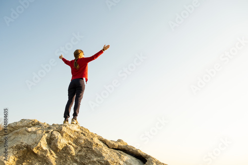 Young woman hiker standing alone on big stone in mountains. Female tourist raising her hands up on high rock in morning nature. Tourism, traveling and healthy lifestyle concept.