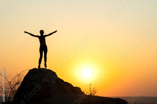Silhouette of a woman hiker standing alone on big stone at sunset in mountains. Female tourist raising her hands up on high rock in evening nature. Tourism, traveling and healthy lifestyle concept.