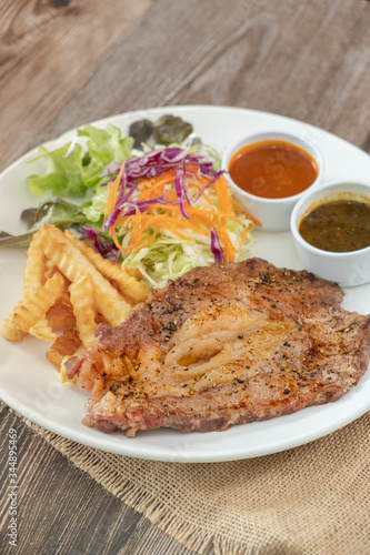 Beef steak with barbecue and paper sauces, side dish salad and french fries served on white plate, wooden table.