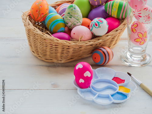 Colorful designs of Easter eggs in straw basket on white wooden surface. Color palette filled with red, yellow and blue color ready to paint with brush for Easter Holidays.