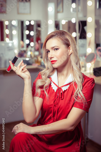 A blonde woman in a vintage red dress in the dressing room sits on a chair and holds her lipstick like a cigarette © Irbis