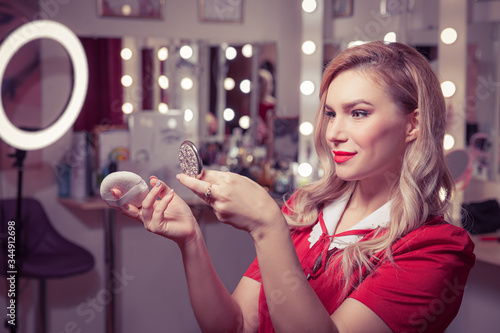 A blonde woman in a vintage red dress in the dressing room looks in a small mirror and adjusts her make